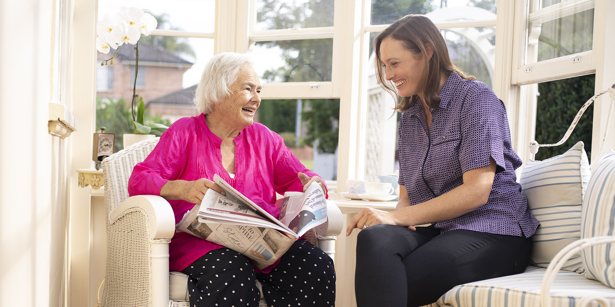 An older woman in a bright pink top sits on a lounge chair while holding a newspaper and chatting with her smiling KinCare home care worker who sits next to her.