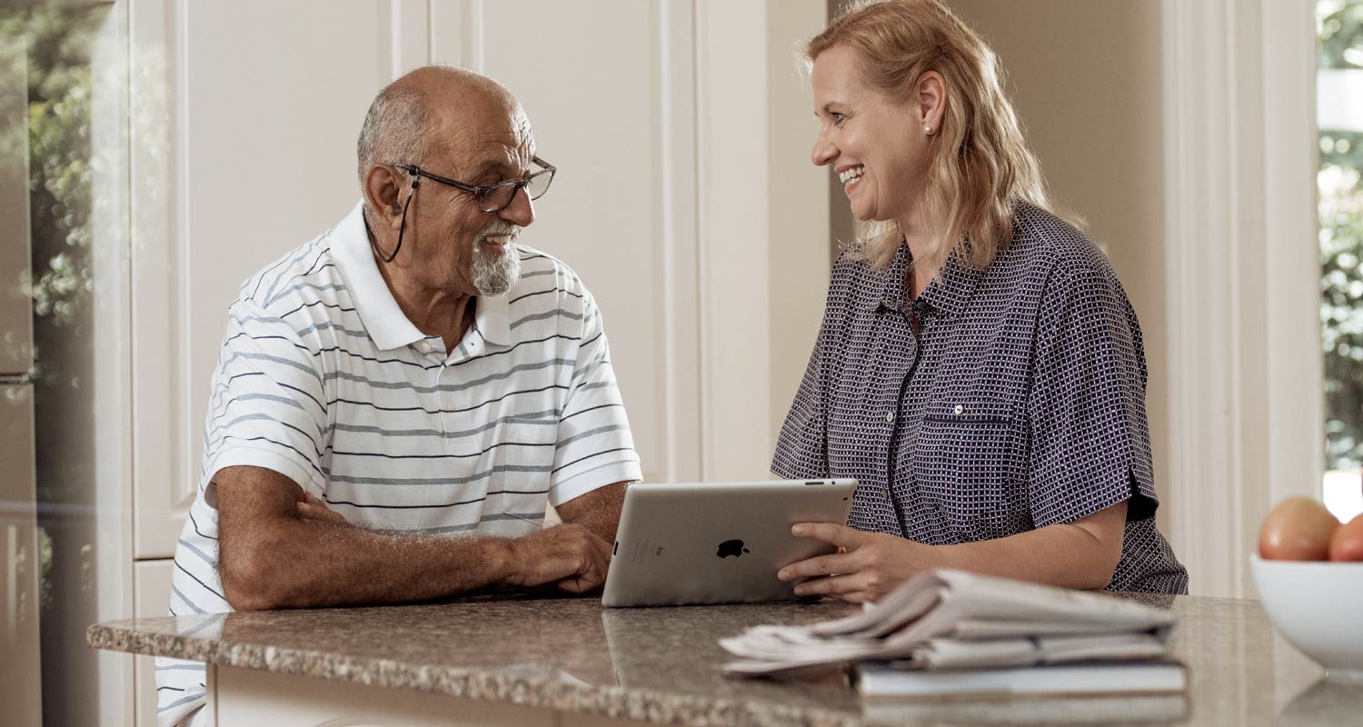 A man in glasses and white striped shirt smiles broadly as he sits at a table with his KinCare home care worker, who is showing him an iPad and smiling.