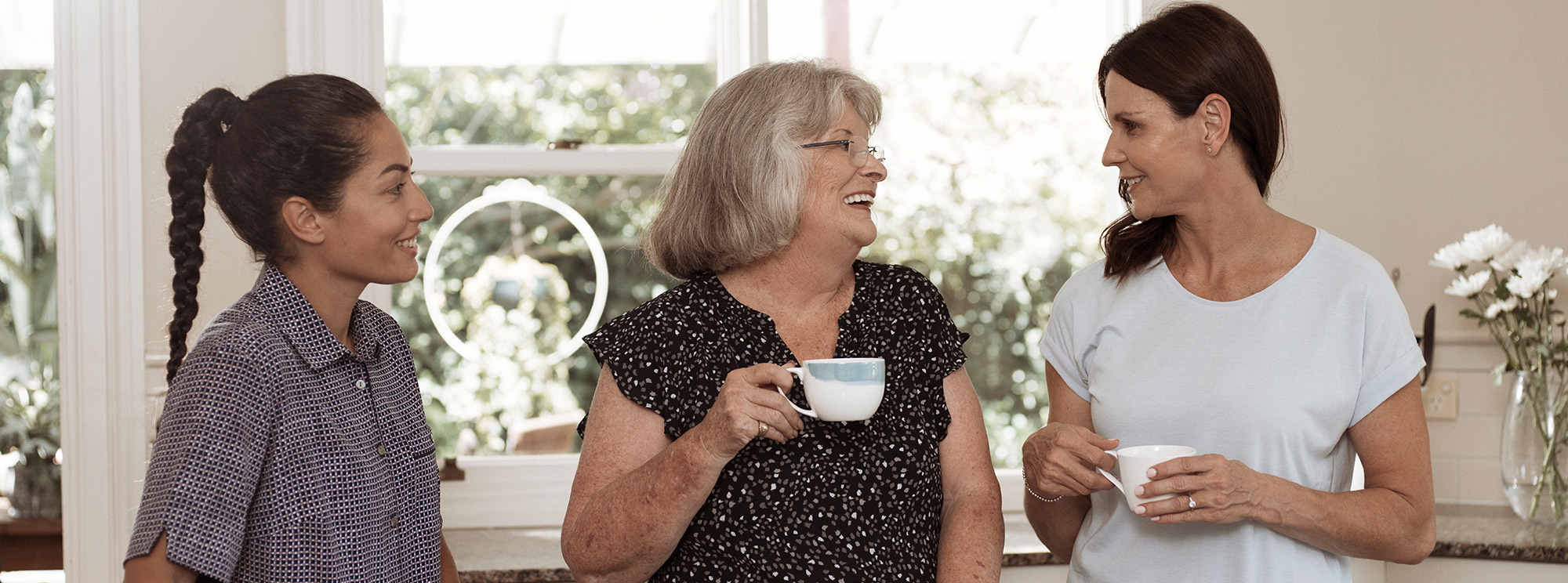 elderly woman and family member chatting with kincare worker around the kitchen bench with tea