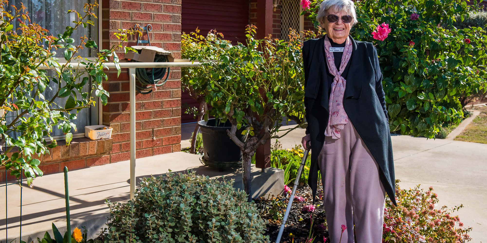 Wilma Mitchell smiling with sunglasses on, standing with her walking stick in her garden