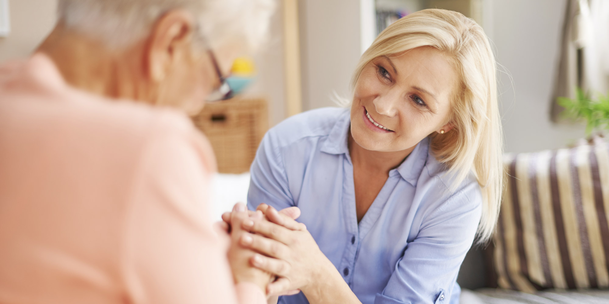 A woman holds an elderly woman's hands between hers and gently smiles.