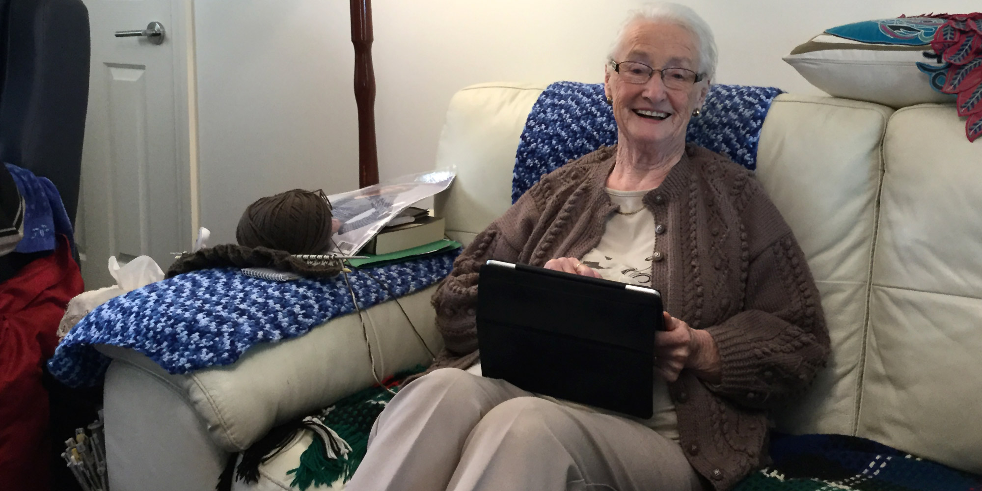 Kincare Customer Mary aged 87 smiling and using her iPad sitting on her sofa at home