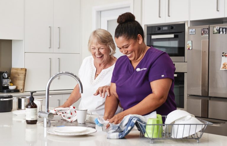 Kincare worker washing the dishes whilst laughing with elderly customer