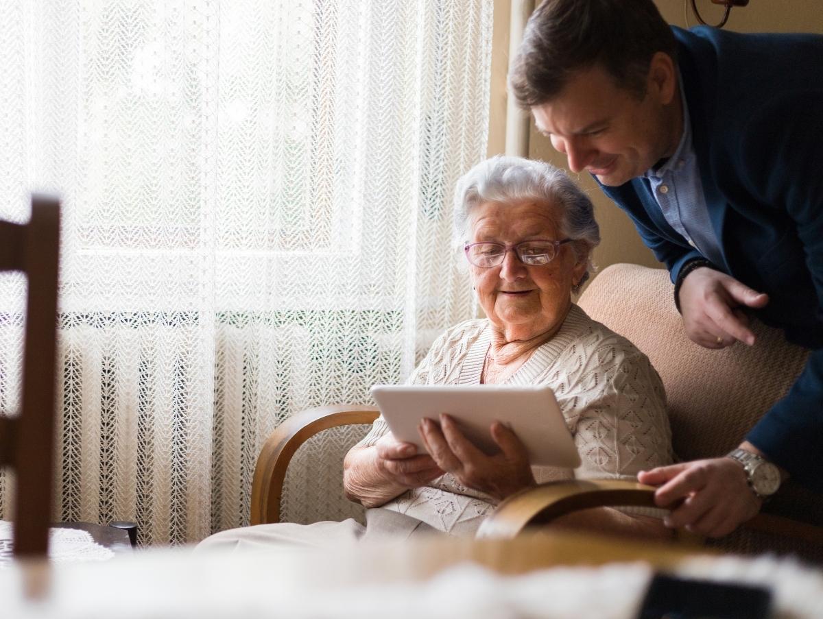 An elderly lady in a comfortable chair, looking at a tablet while a mddle-aged man rests his arm on the couch behind her.
