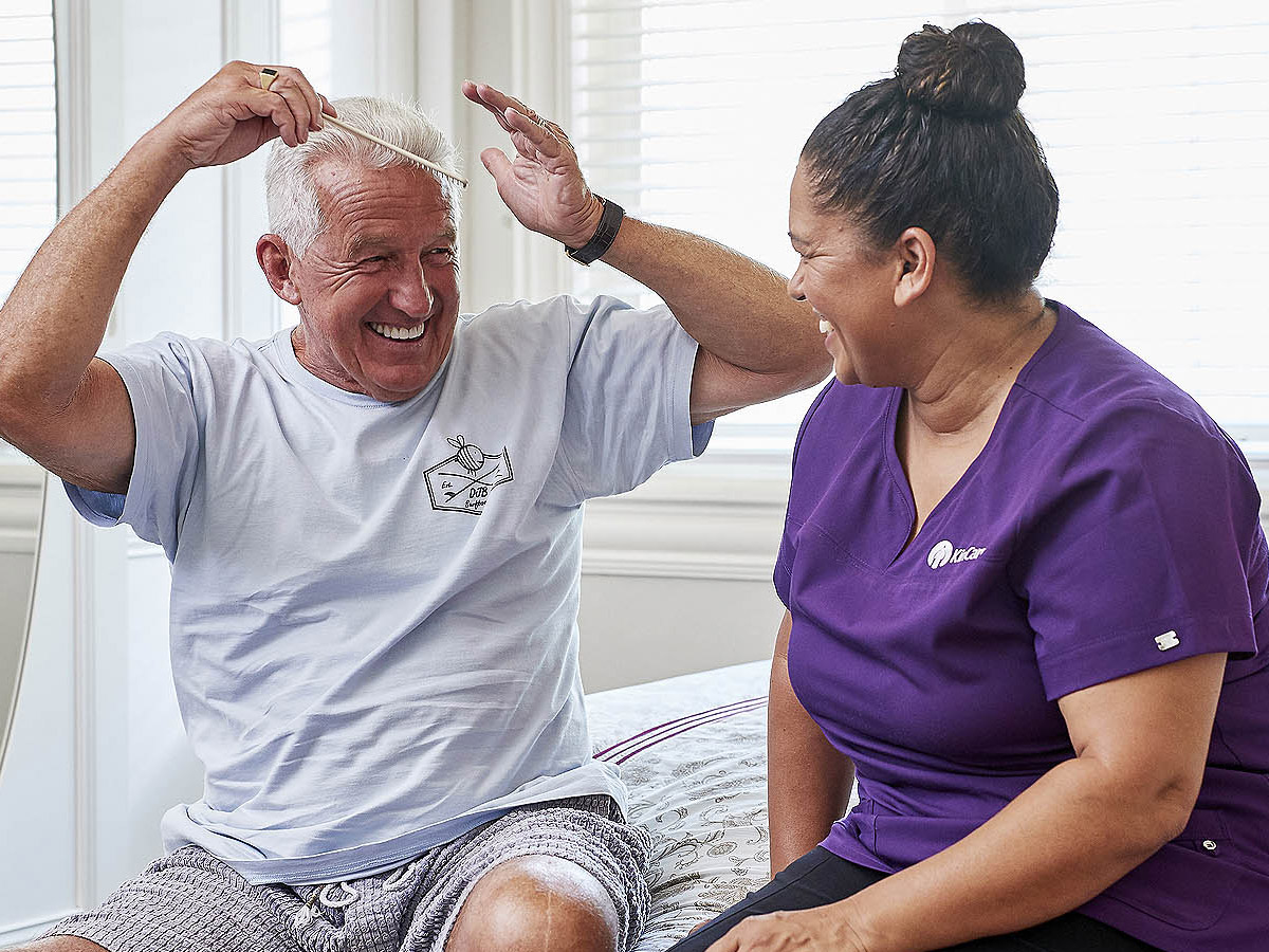 A white-haired man combs his hair and laughs with his KinCare home care worker.