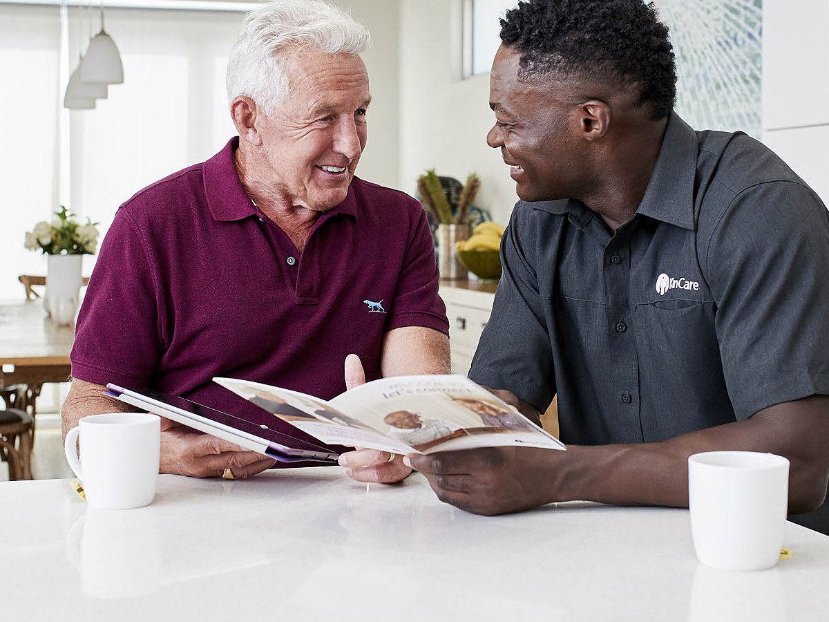 A white-haired man and his KinCare Home Care Worker chatting at the kitchen table with brochures.