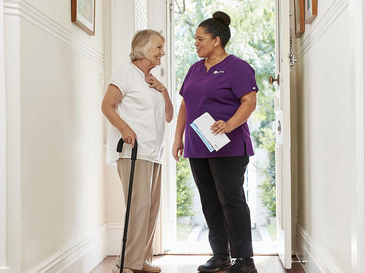 A lady supporting herself inside her front door with a cane and chatting with her KinCare Home Care Worker.