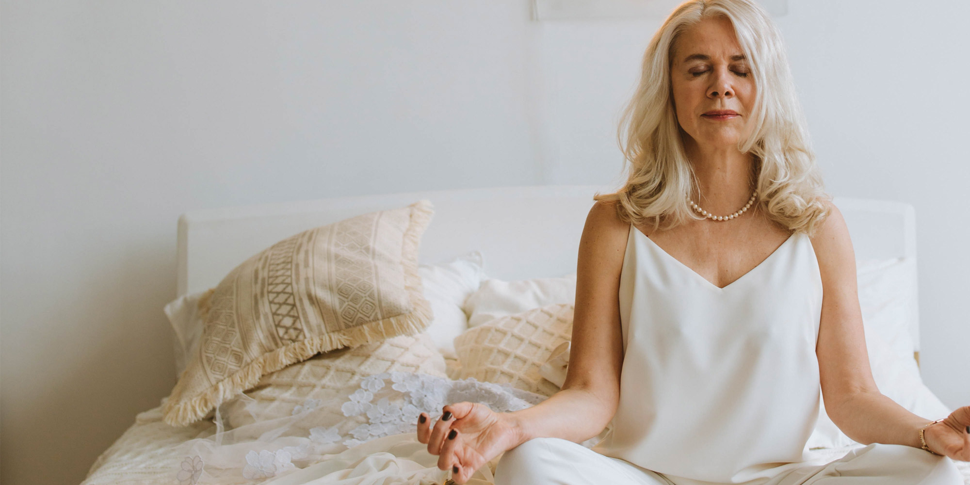 A meditating woman sits cross-legged on her bed with her hands resting on her knees.